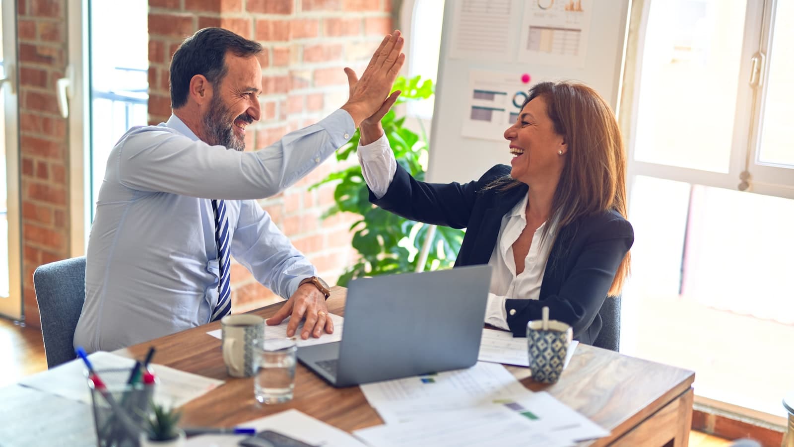 Two colleagues celebrating a milestone with a high five at a desk with a laptop and paperwork