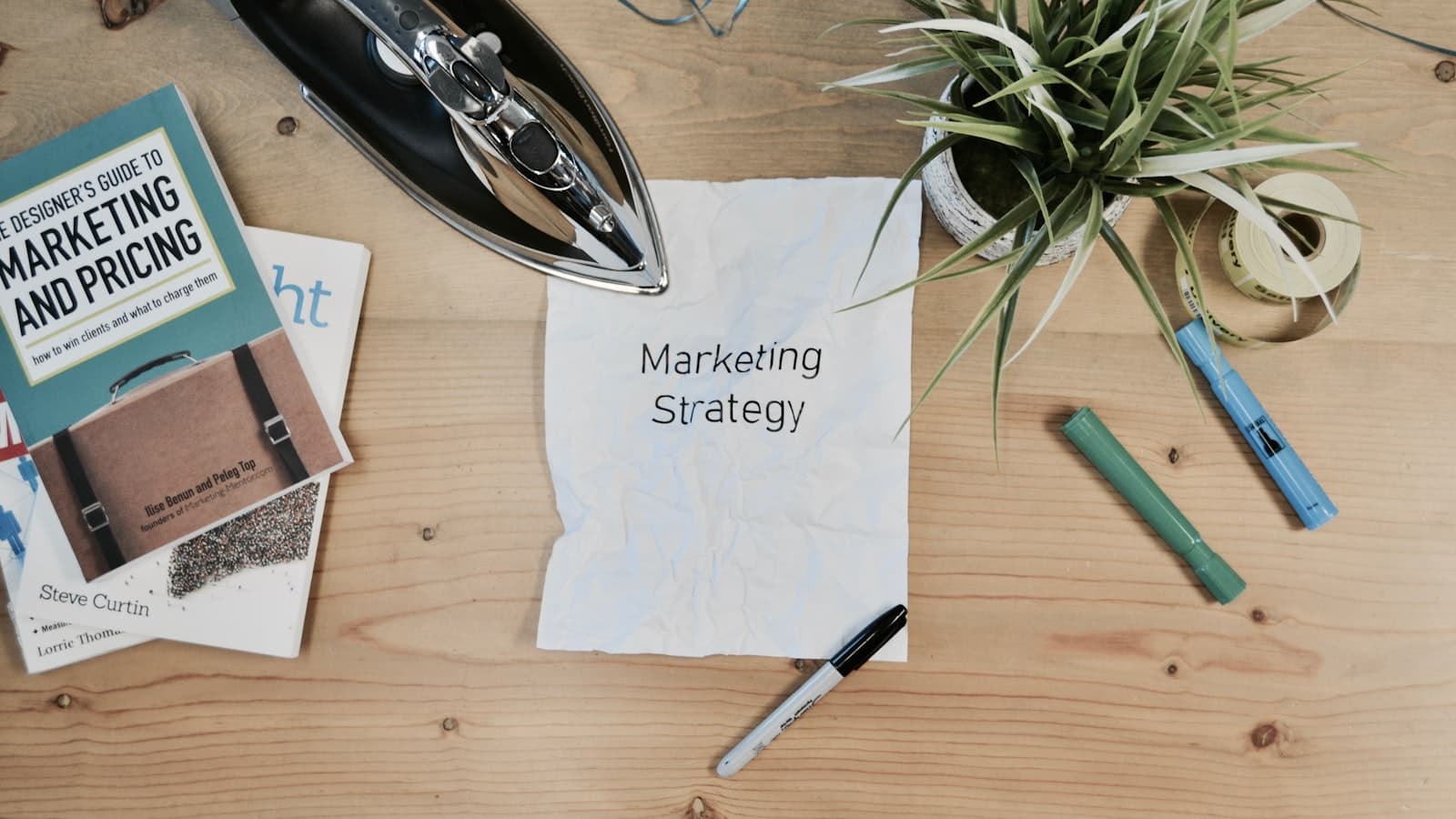 Desk flat lay with a marketing strategy note, a marketing book, pens and a small plant